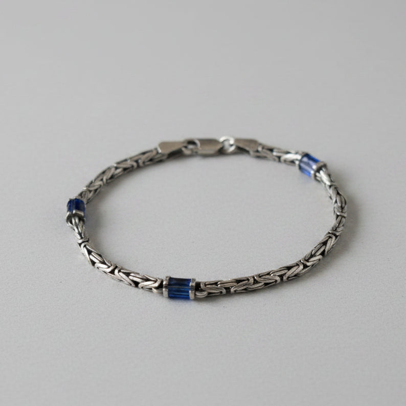 A silver-toned metal chain bracelet with small dark blue rectangular accents, displayed on a black background.Product DetailsStyle: Interlinked chain links with a polished finish.Accents: Small, dark blue rectangular components embedded symmetrically along the band.Clasp: Traditional lobster claw closure.Background: The image currently displays the bracelet resting on a plain, light grey surface.