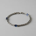 A silver-toned metal chain bracelet with small dark blue rectangular accents, displayed on a black background.Product DetailsStyle: Interlinked chain links with a polished finish.Accents: Small, dark blue rectangular components embedded symmetrically along the band.Clasp: Traditional lobster claw closure.Background: The image currently displays the bracelet resting on a plain, light grey surface.